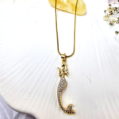 Close-up of gold-plated mermaid pendant necklace on white platter showing pavé crystal tail and snake chain.