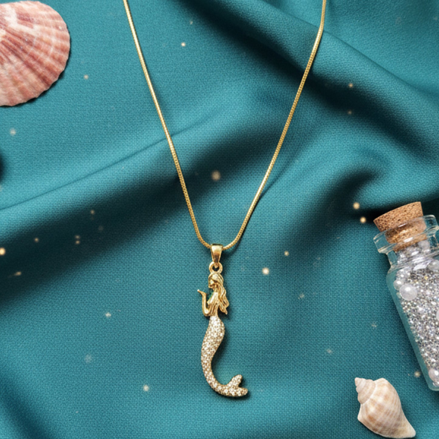 Gold mermaid charm necklace styled with seashells on a teal blue silk background, enhancing the ocean theme.