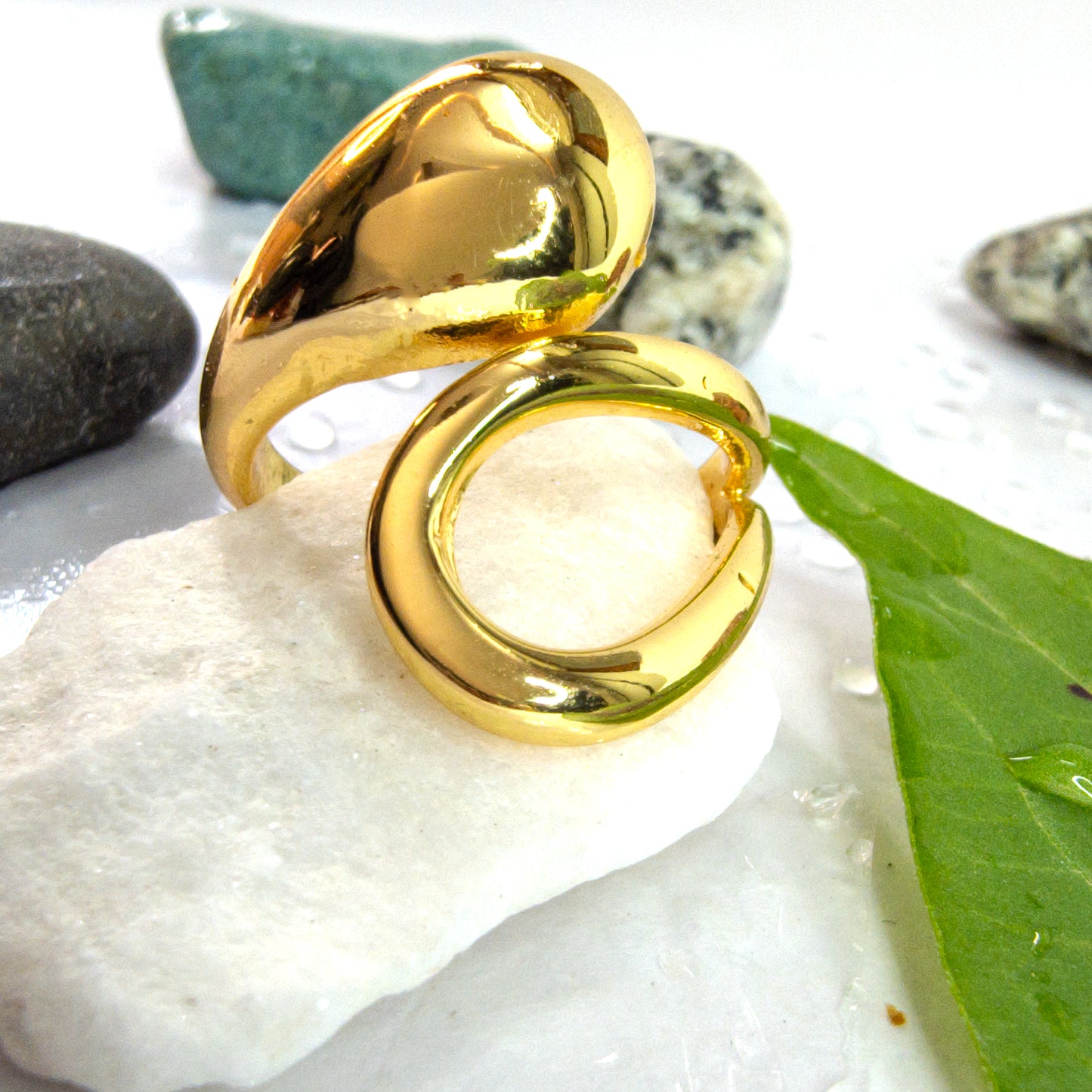 Gold molten drop ring displayed on white stone with green leaf and water droplets, elegant jewelry close-up.