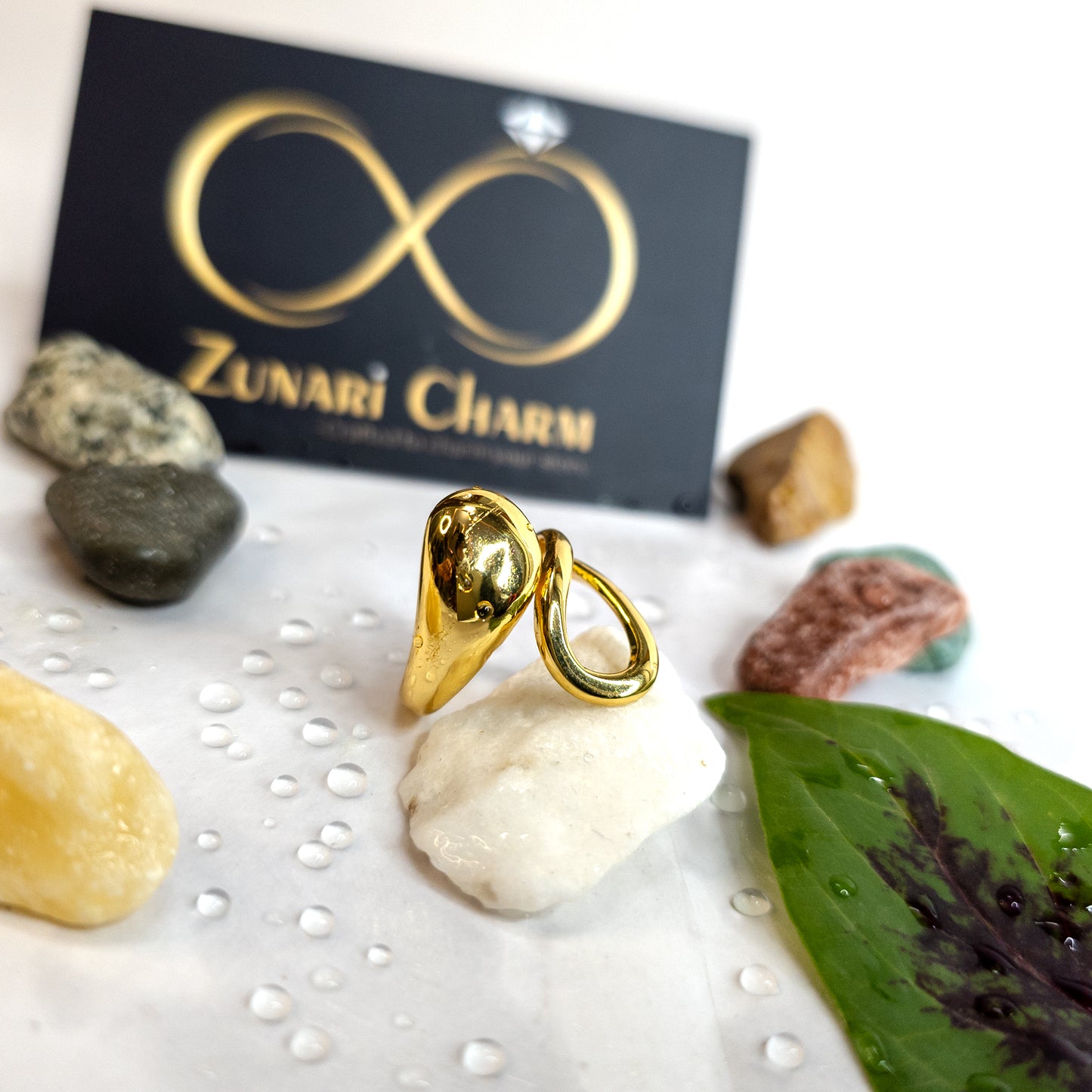 Gold molten drop statement ring on display with natural stones, green leaf, and Zunari Charm branding.