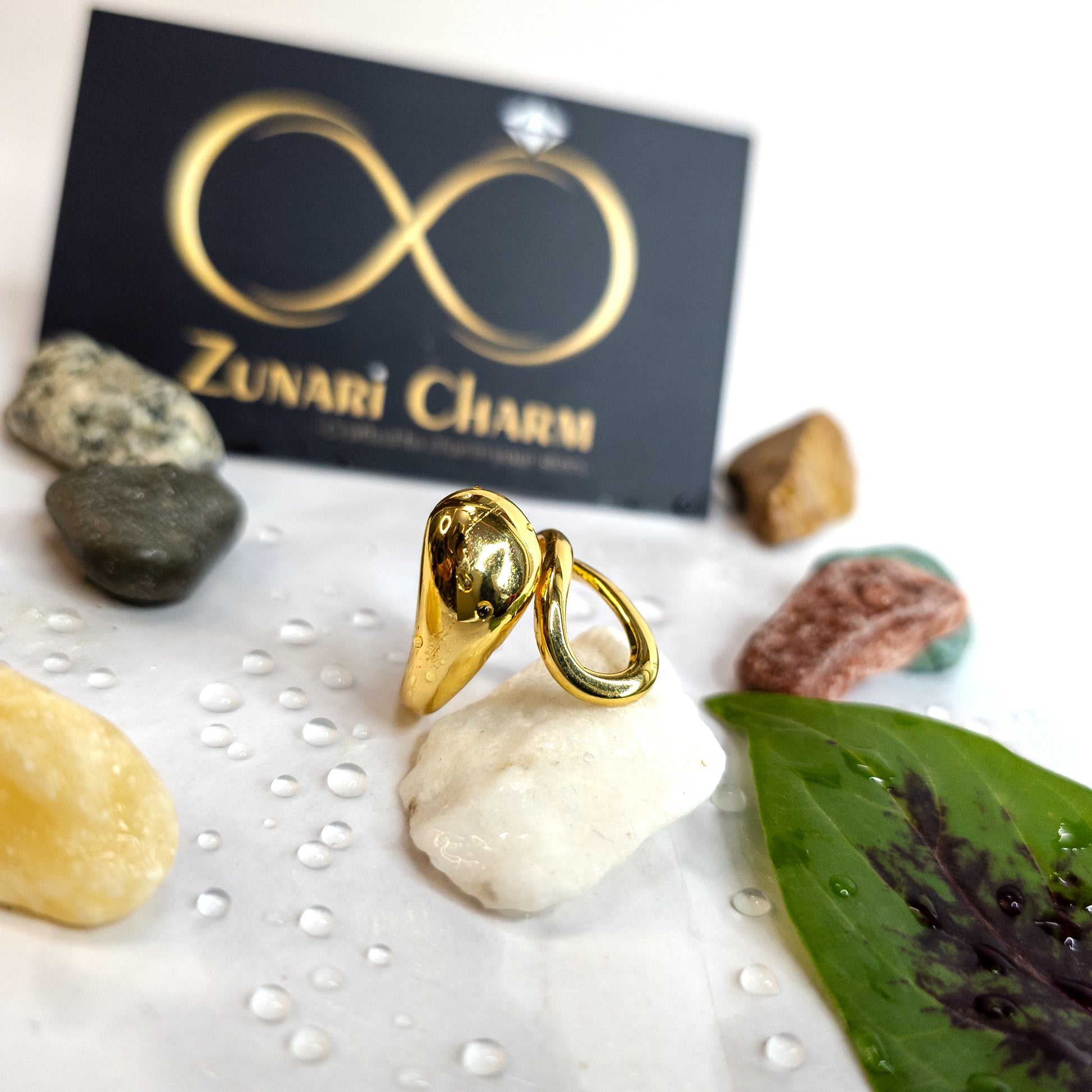 Gold molten drop statement ring on display with natural stones, green leaf, and Zunari Charm branding.