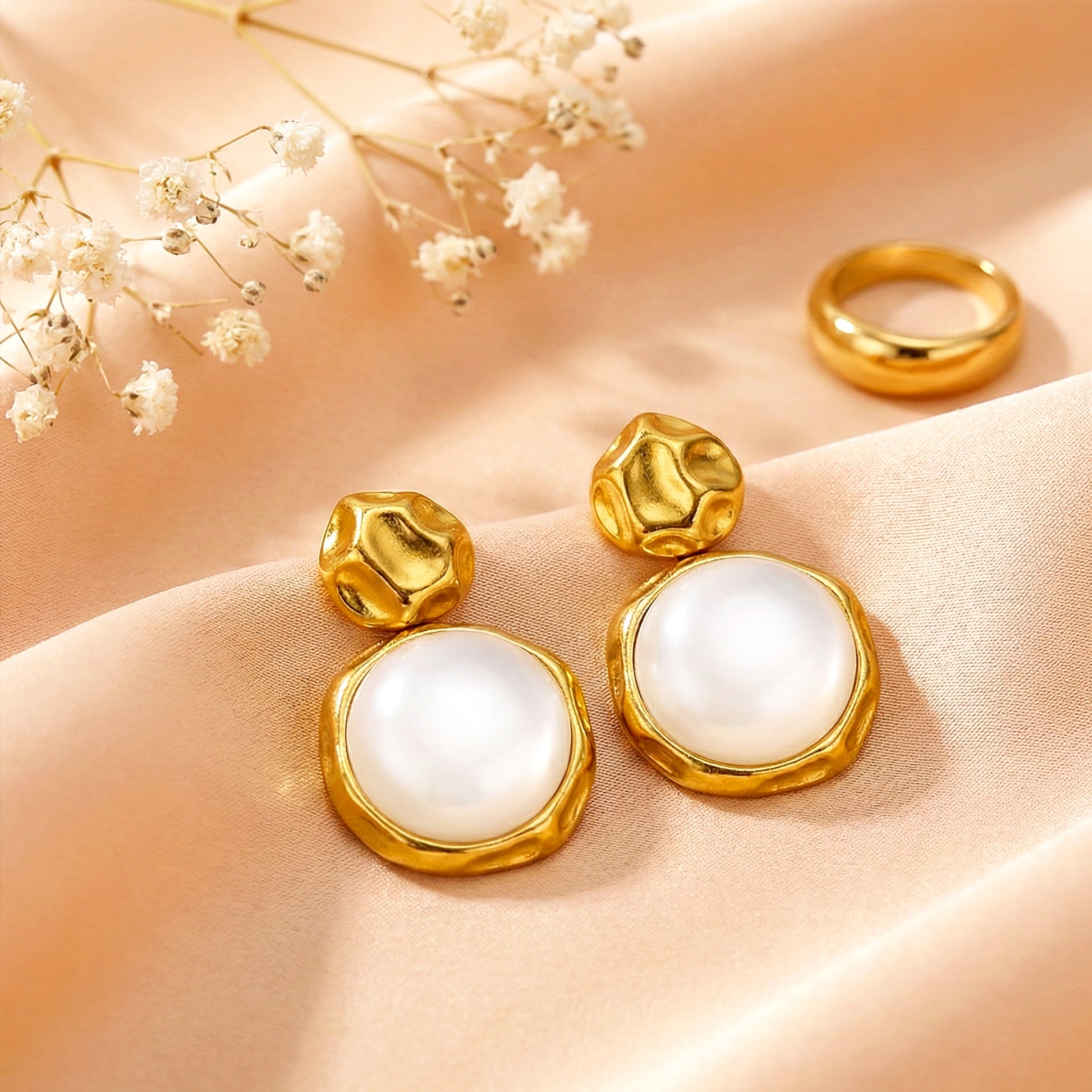 Aesthetic flat lay of moon drop earrings on a peach silk cloth next to a gold ring and dried white flowers.