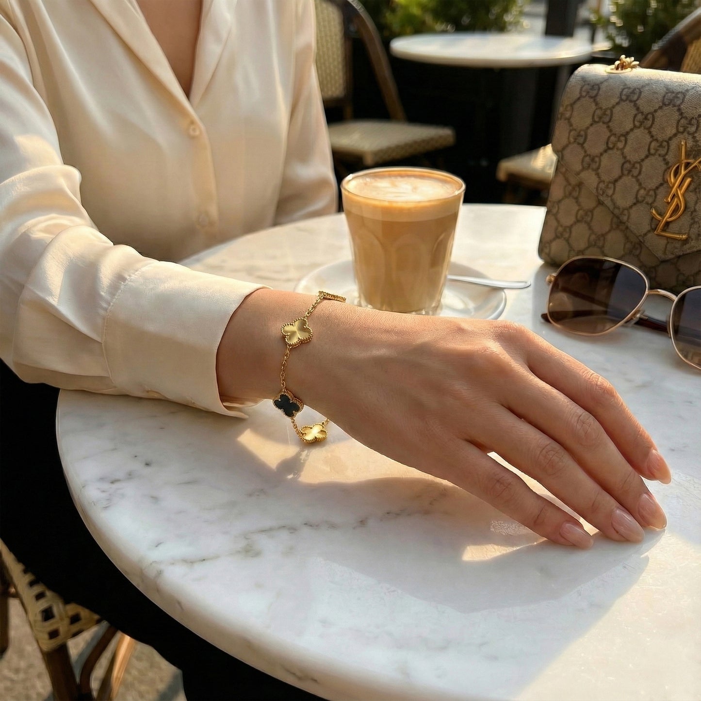 Woman wearing luxury black and gold clover bracelet at a coffee shop with designer accessories.