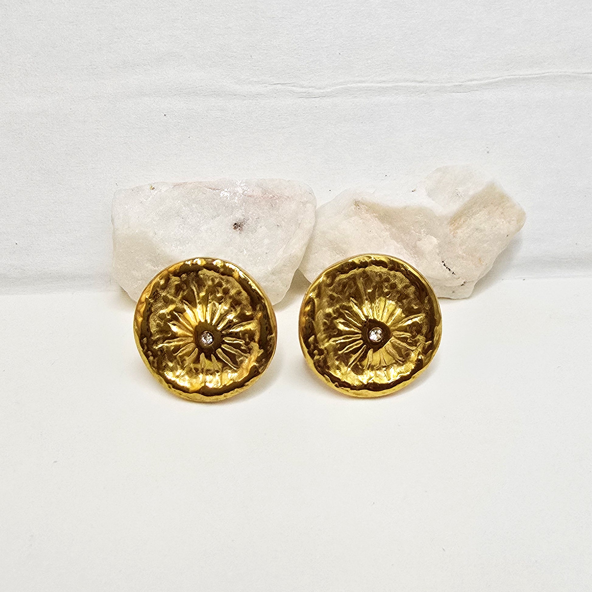 A pair of high-polish gold coin earrings with an etched sunburst pattern and tiny central stone on white decorative rocks.