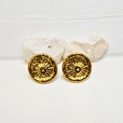 A pair of high-polish gold coin earrings with an etched sunburst pattern and tiny central stone on white decorative rocks.