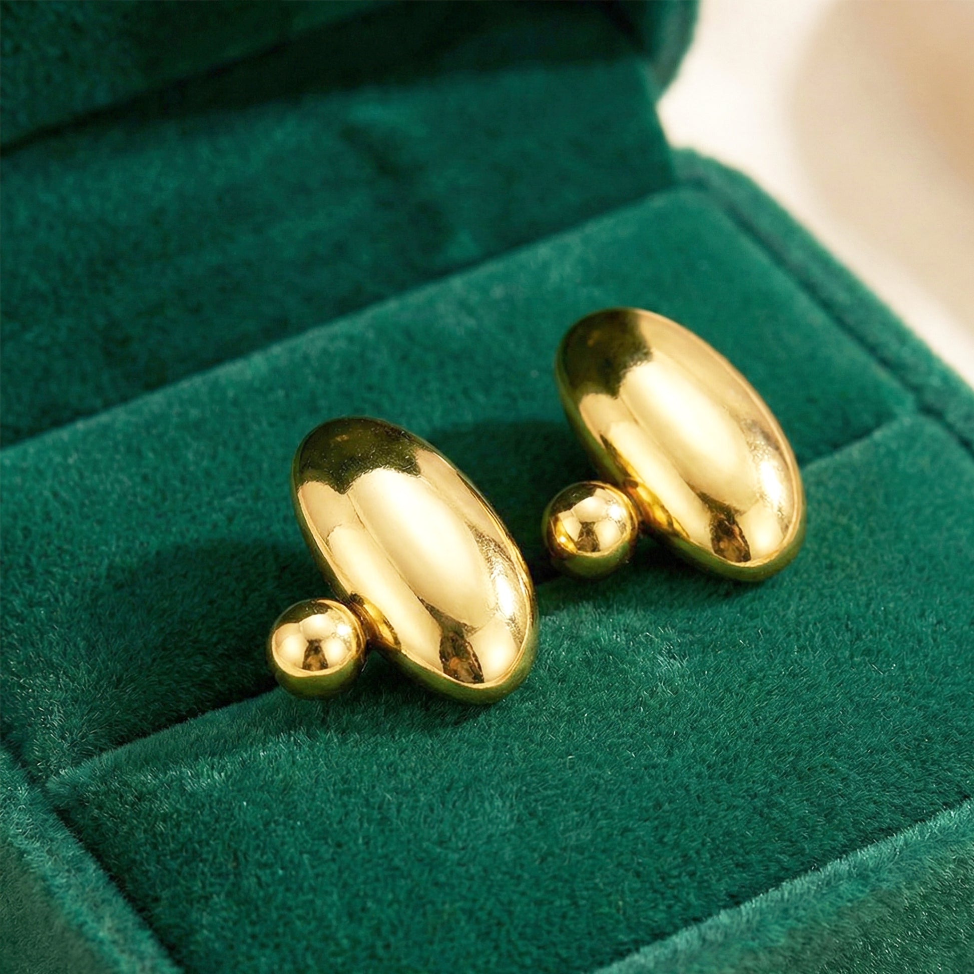 Aesthetic photography of gold pebble studs nestled in a green velvet jewelry box under warm lighting.