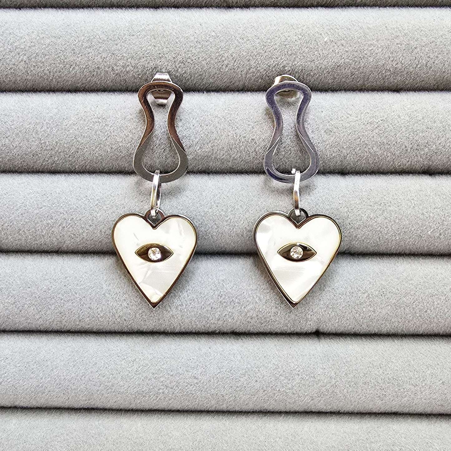 A pair of silver-toned Prism Eye earrings with heart-shaped mother-of-pearl inlays on a grey velvet tray.