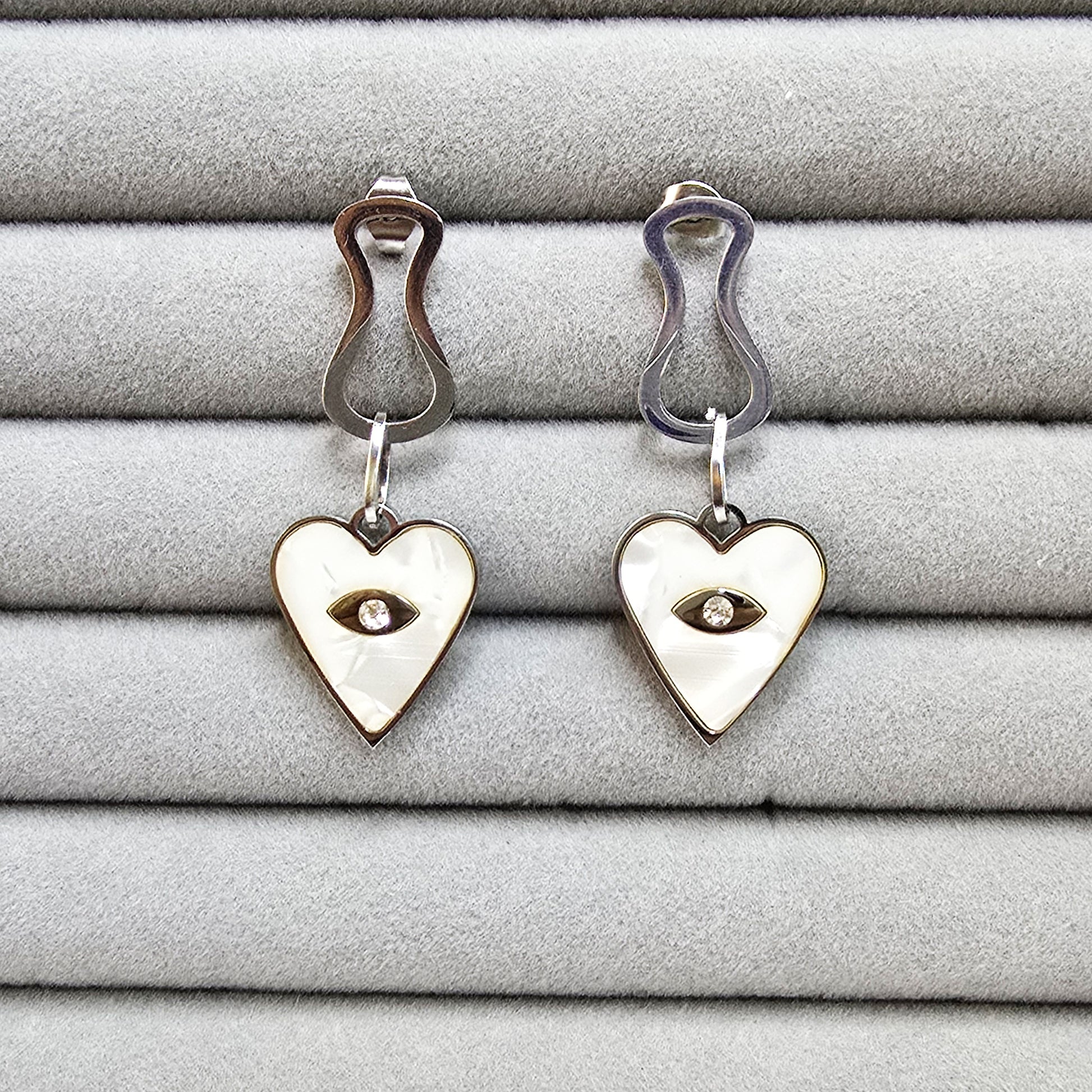 A pair of silver-toned Prism Eye earrings with heart-shaped mother-of-pearl inlays on a grey velvet tray.