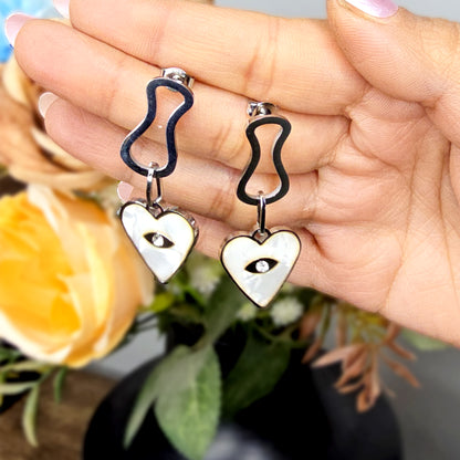 Hand holding silver heart dangle earrings to show the protective eye symbol and iridescent mother-of-pearl finish.