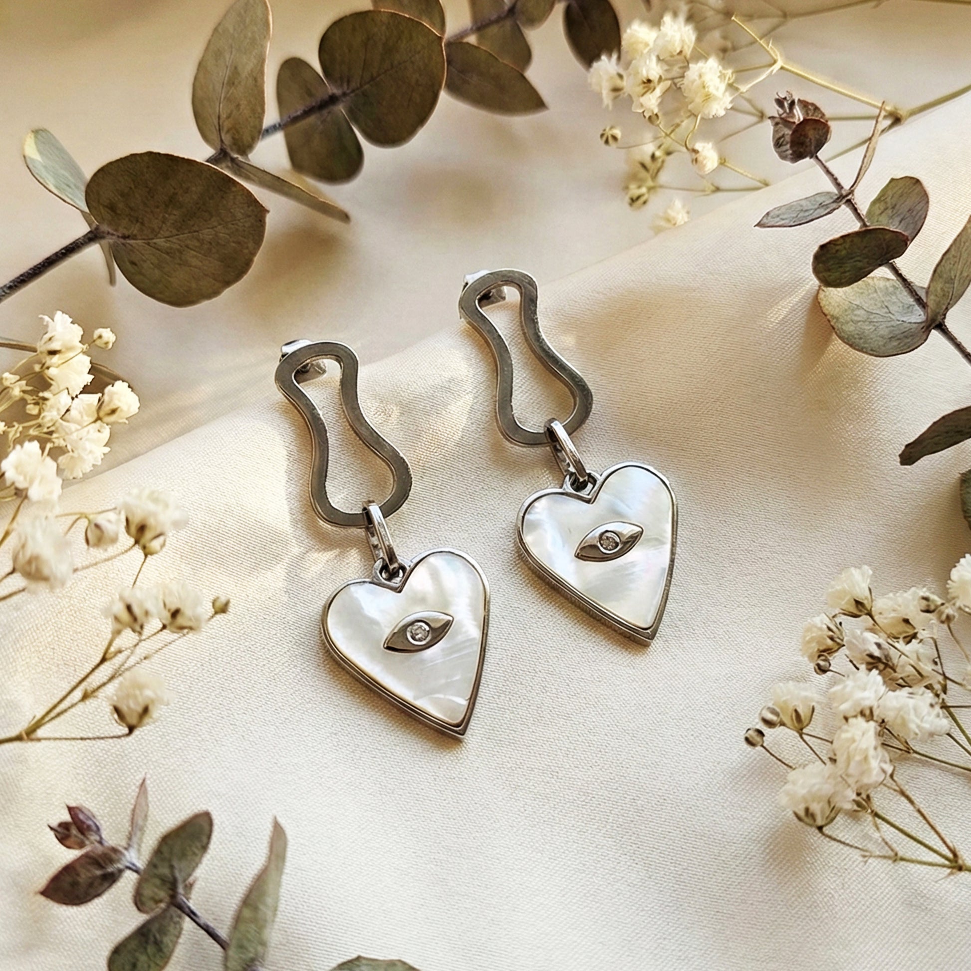 Aesthetic photography of silver heart dangles on a cream fabric next to dried eucalyptus and white flowers.