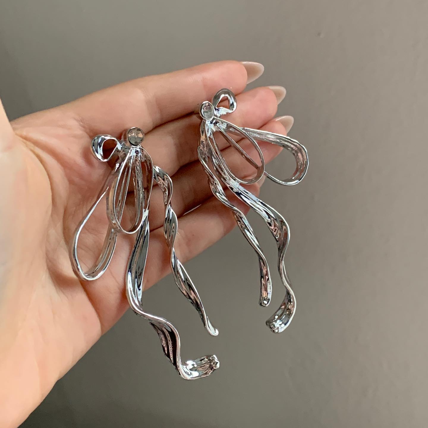 Hand holding both exaggerated silver bow earrings with visible twisted design
