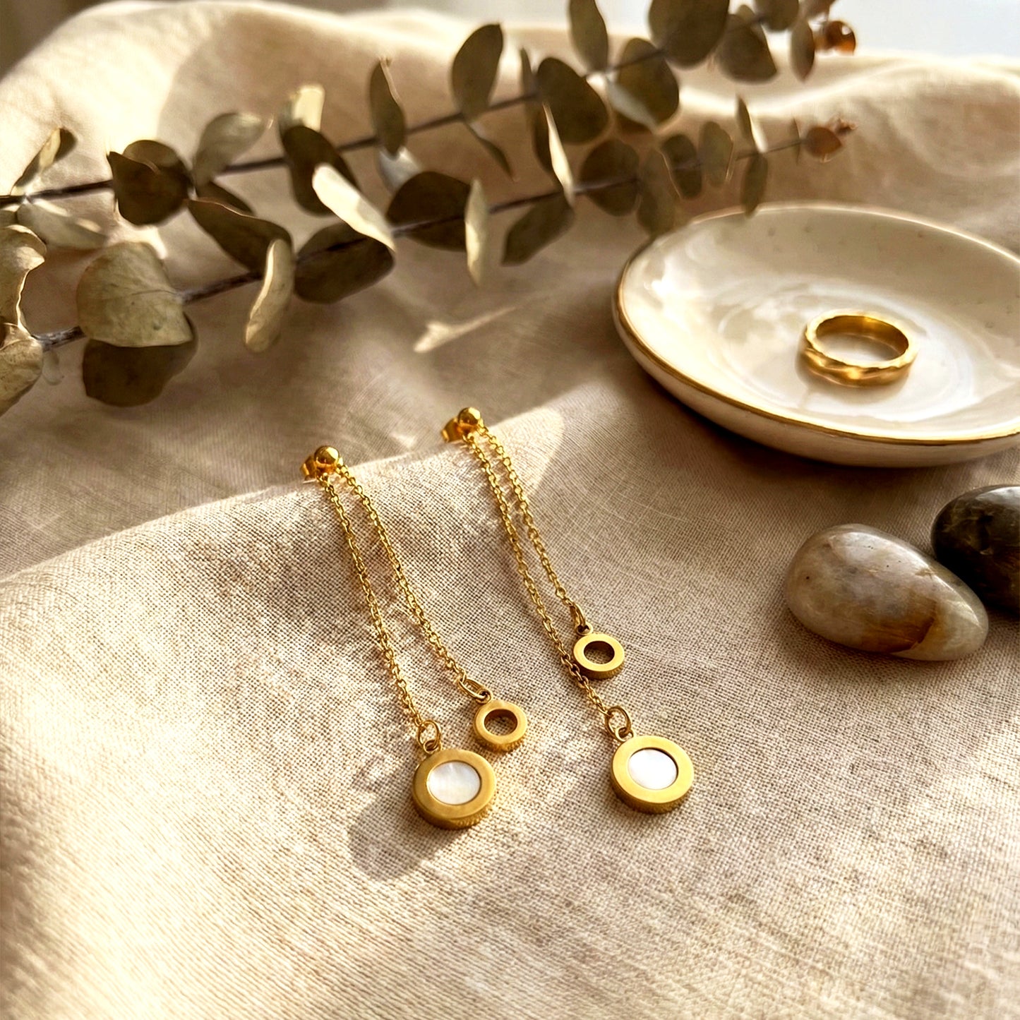 Aesthetic photography of gold Slide Drop earrings on beige linen cloth next to a gold ring and eucalyptus.