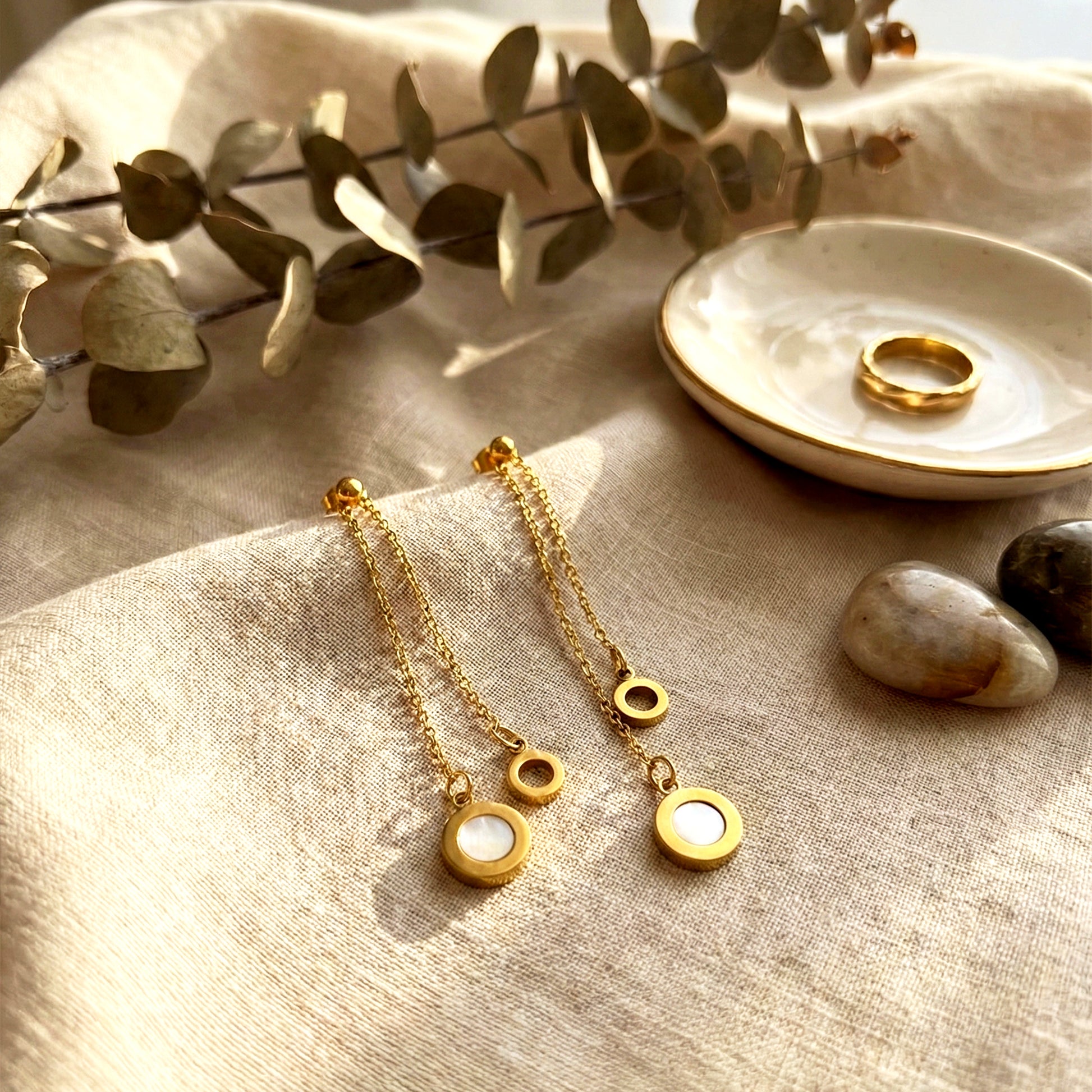 Aesthetic photography of gold Slide Drop earrings on beige linen cloth next to a gold ring and eucalyptus.
