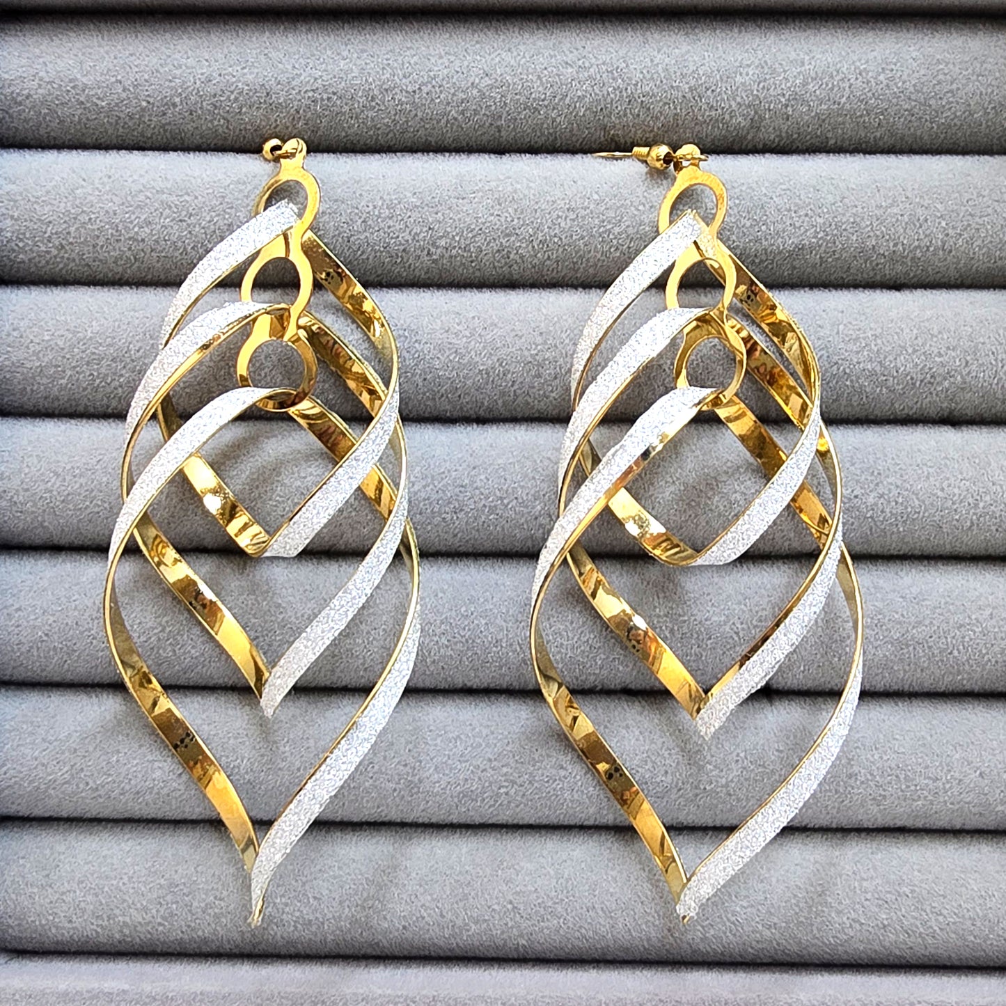 A pair of large dual-tone gold and silver glitter leaf-shaped dangling earrings on a grey velvet tray.