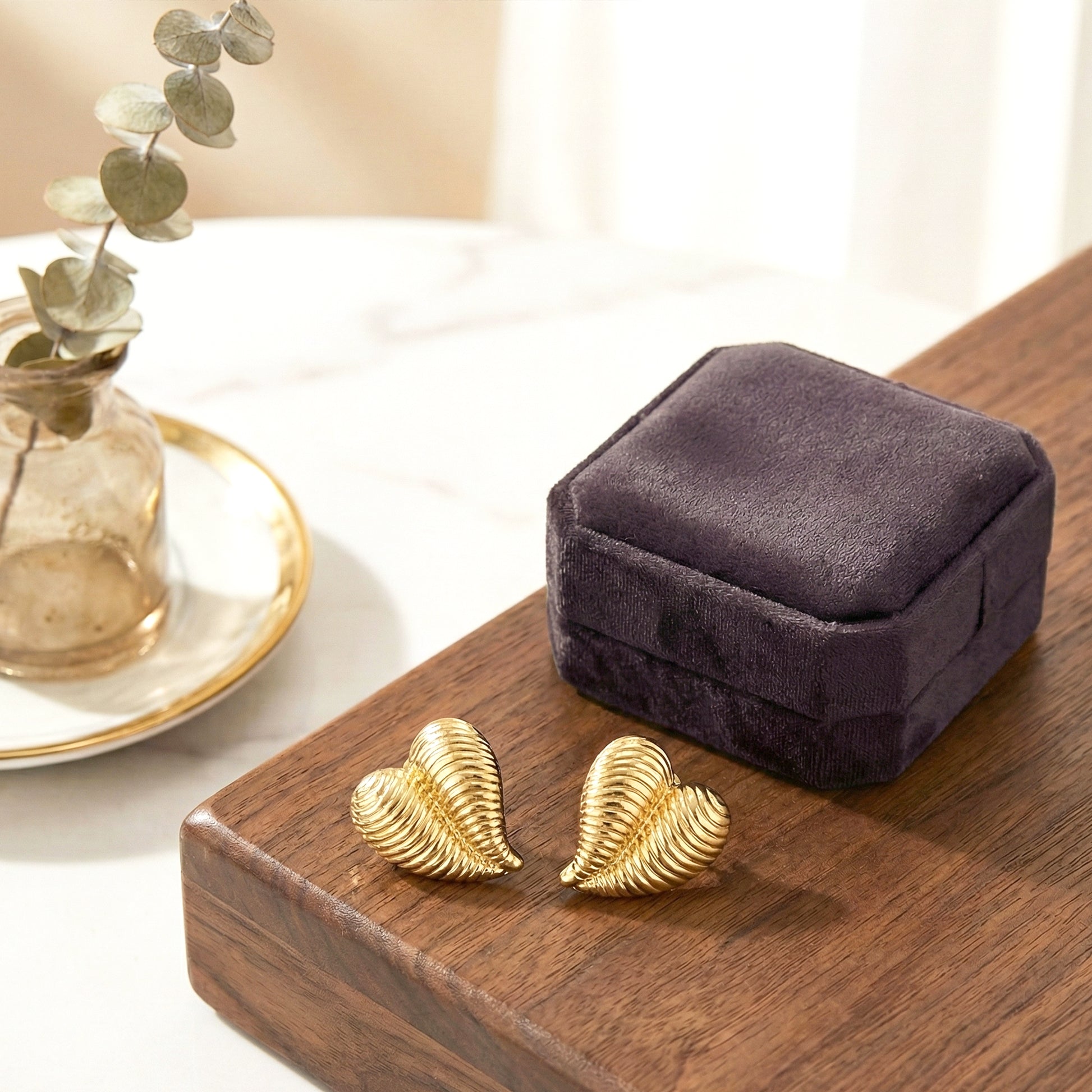 Aesthetic photography of gold ribbed heart earrings placed next to a dark purple velvet jewelry box on a wooden surface.
