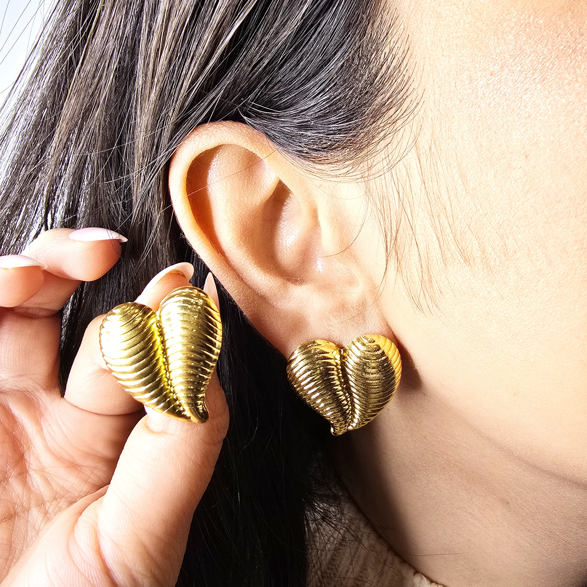 Macro shot of a model wearing a large gold ribbed heart stud earring, showcasing the scale and textured finish.
