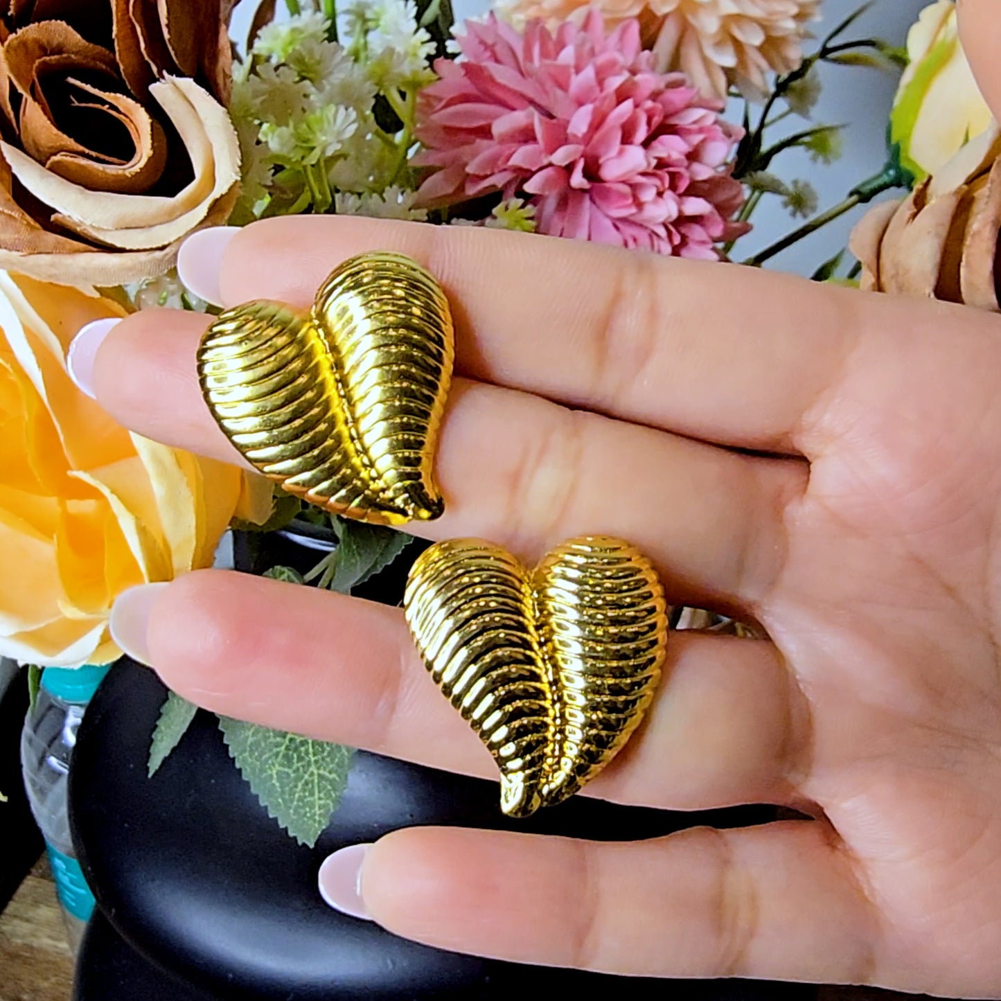 Hand holding two high-shine gold striped heart earrings to demonstrate the reflective ribbed detail and metallic finish.
