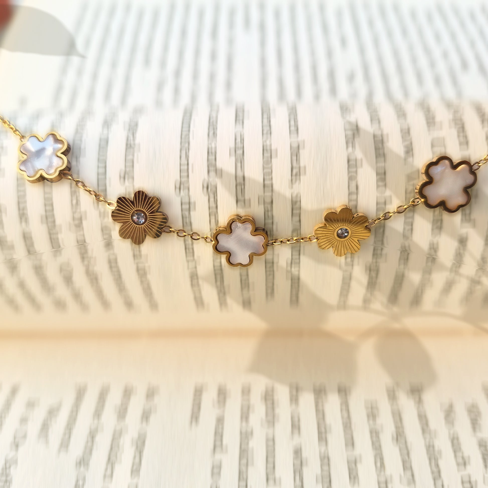 Gold bracelet with alternating white pearly clovers and textured sunburst charms draped on a book.