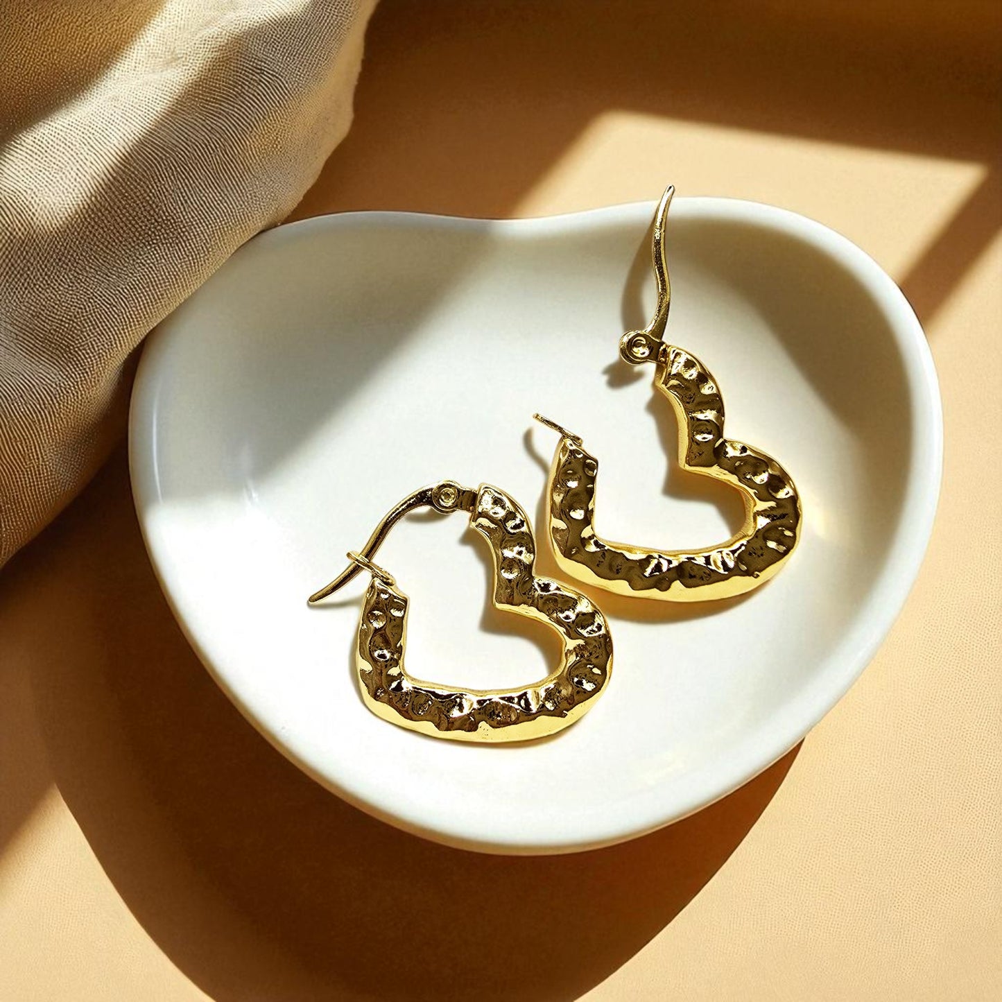 Gold textured heart hoop earrings placed inside a white dish with warm lighting, highlighting hammered gold texture.