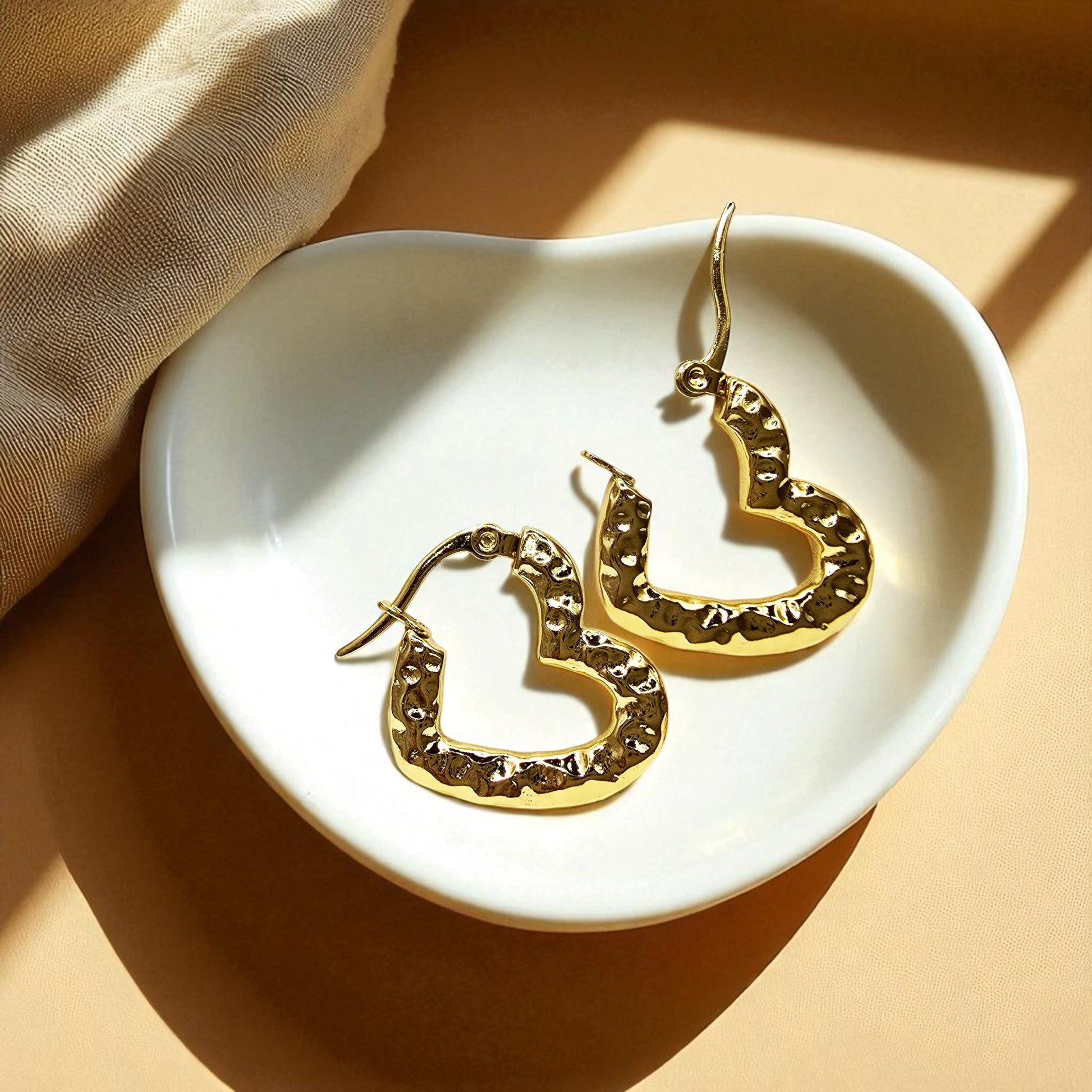Gold textured heart hoop earrings placed inside a white dish with warm lighting, highlighting hammered gold texture.