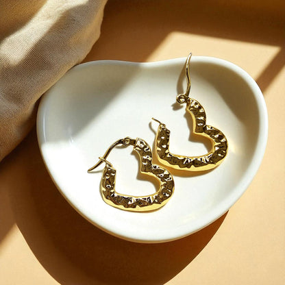 Gold textured heart hoop earrings placed inside a white dish with warm lighting, highlighting hammered gold texture.