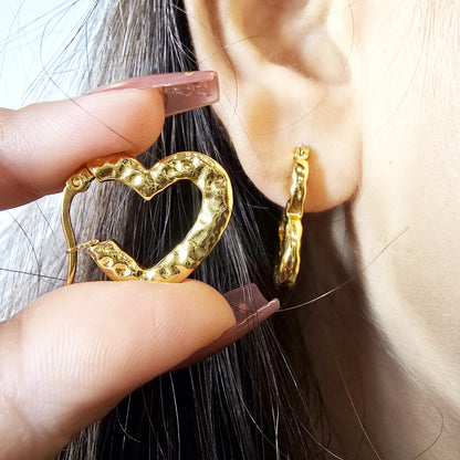 Close-up of model wearing gold textured heart hoop earring, with hand holding the second earring near the ear.