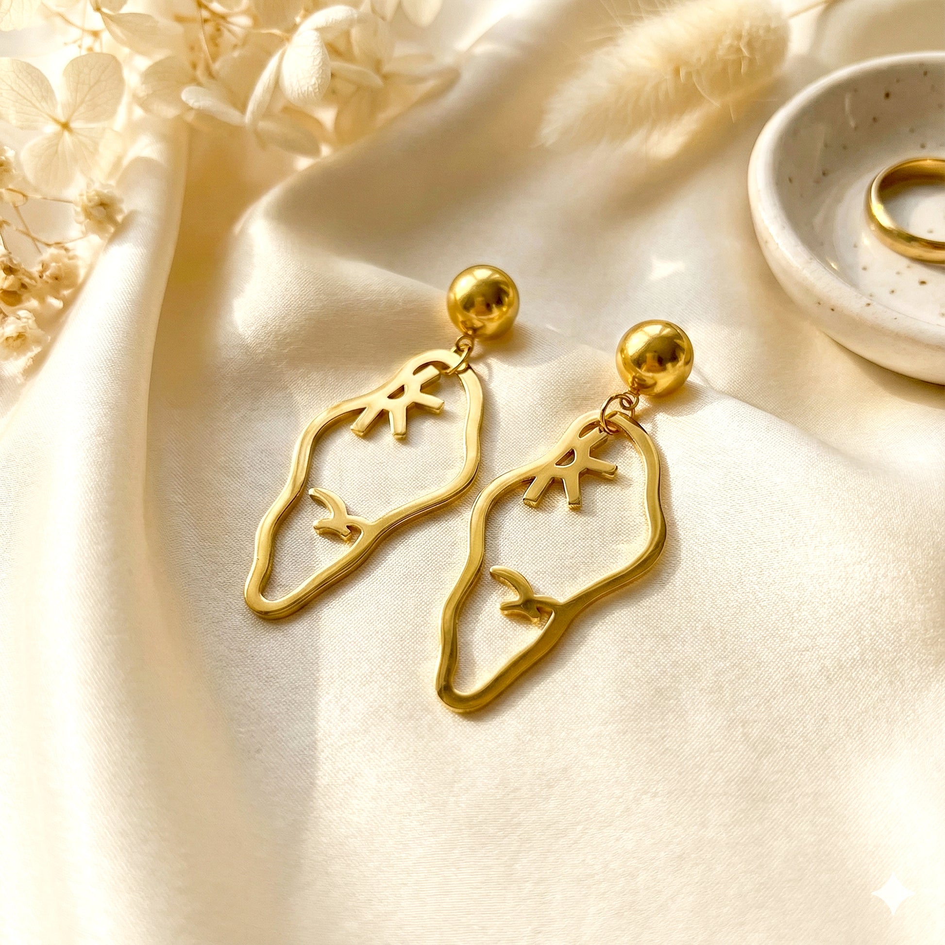 Aesthetic photography of gold Vertex earrings on a cream silk cloth with a small gold dish and dried flowers.