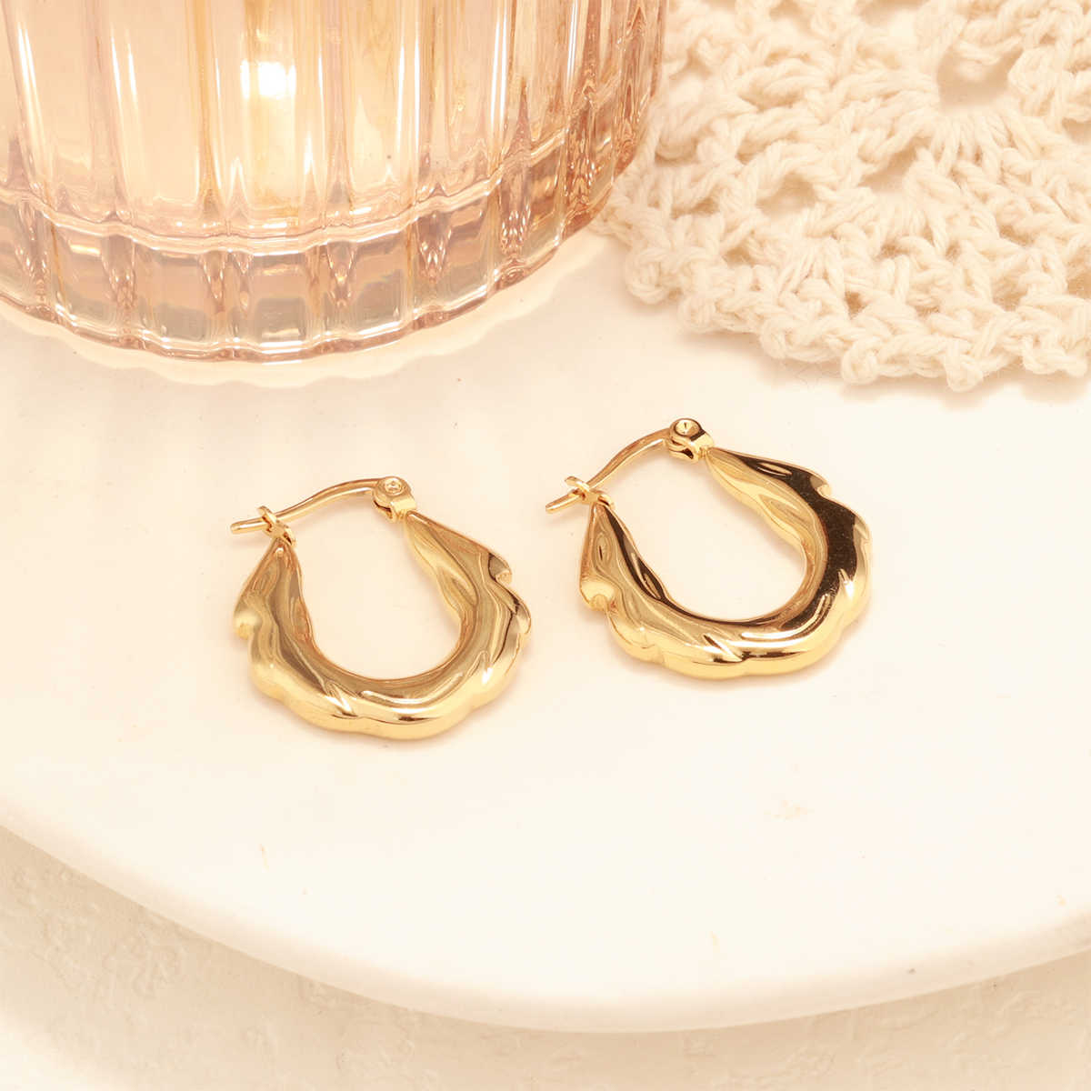 One gold zigzag hoop earring propped against a blush-toned glass with crochet detailing in background.