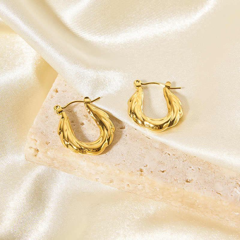 Sculptural gold zigzag hoop earrings styled on a textured beige stone tile background.