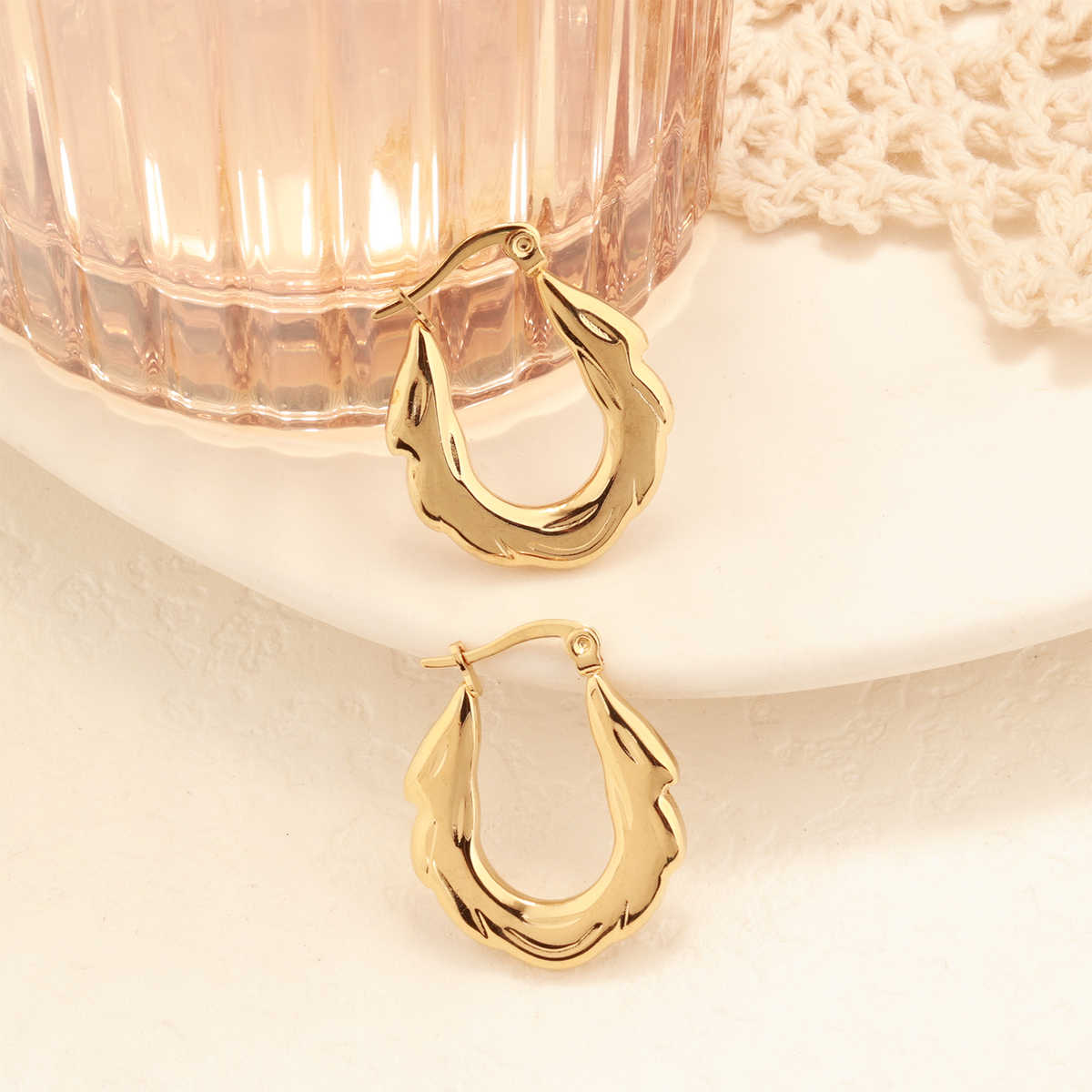 Gold zigzag hoop earrings placed beside a textured glass tumbler and lace fabric on a white ceramic plate
