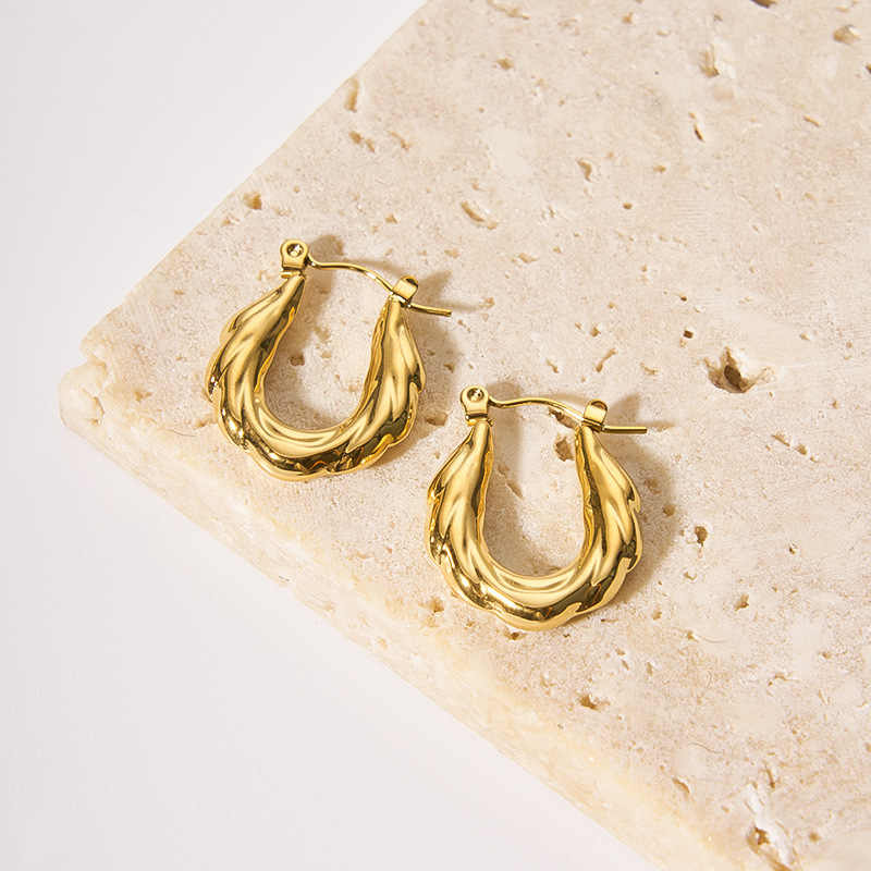 Sculptural gold zigzag hoop earrings styled on a textured beige stone tile background.