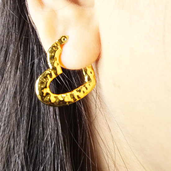 Close-up of a model wearing gold textured heart hoop earring, hand gently touching the earring to highlight its hammered design.